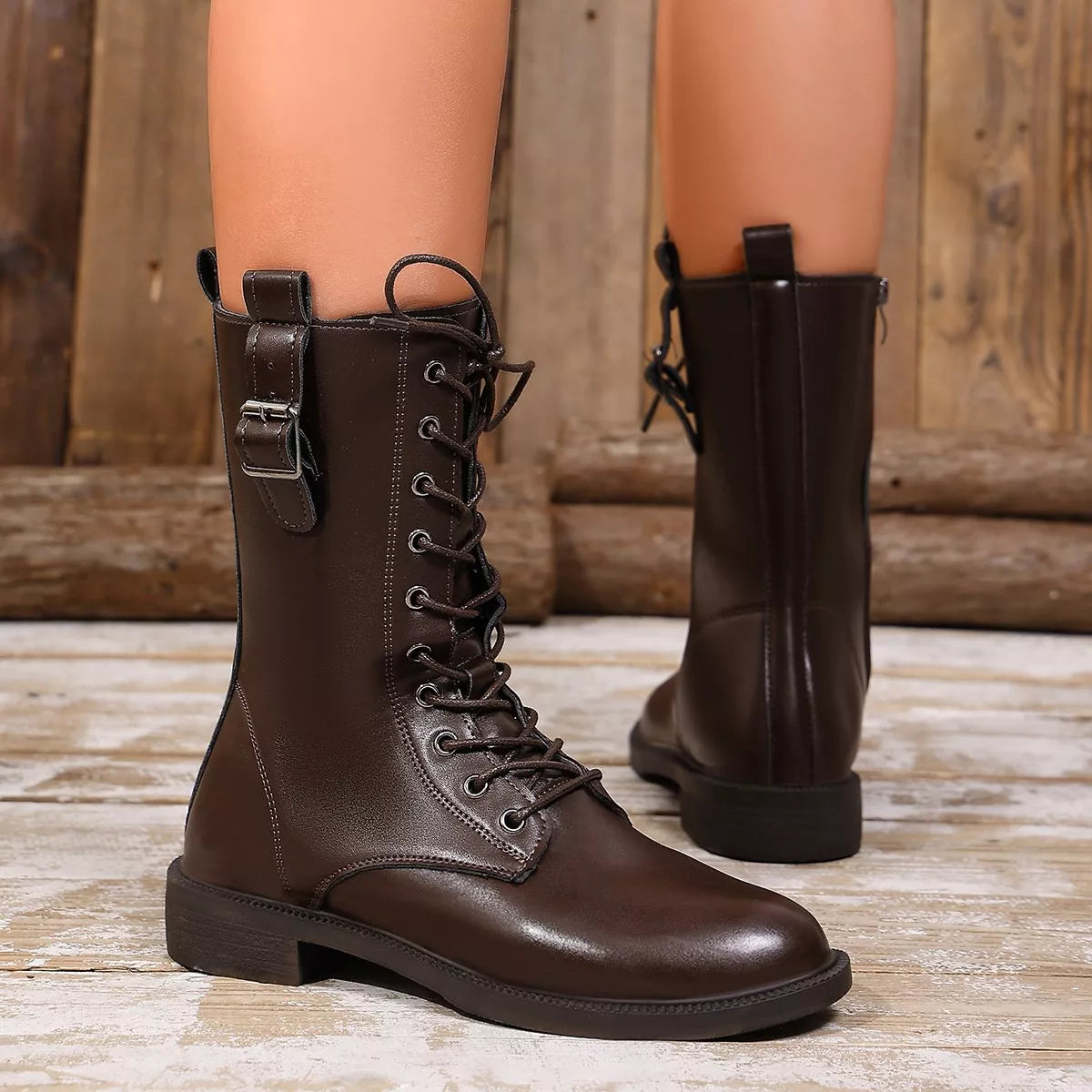 NEWYORKLISTED SHOE COLLECTION: A pair of dark brown leather lace-up combat boots worn on a mannequin or model's legs, shown against a rustic wooden background.