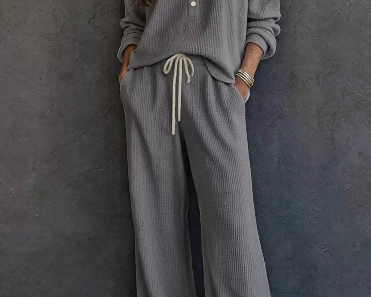 NEWYORKLISTED STYLE & COMFORT COLLECTION: A woman wearing a matching gray waffle-knit lounge set with a long-sleeve henley top and wide-leg pants with a drawstring waist, standing against a dark textured wall.