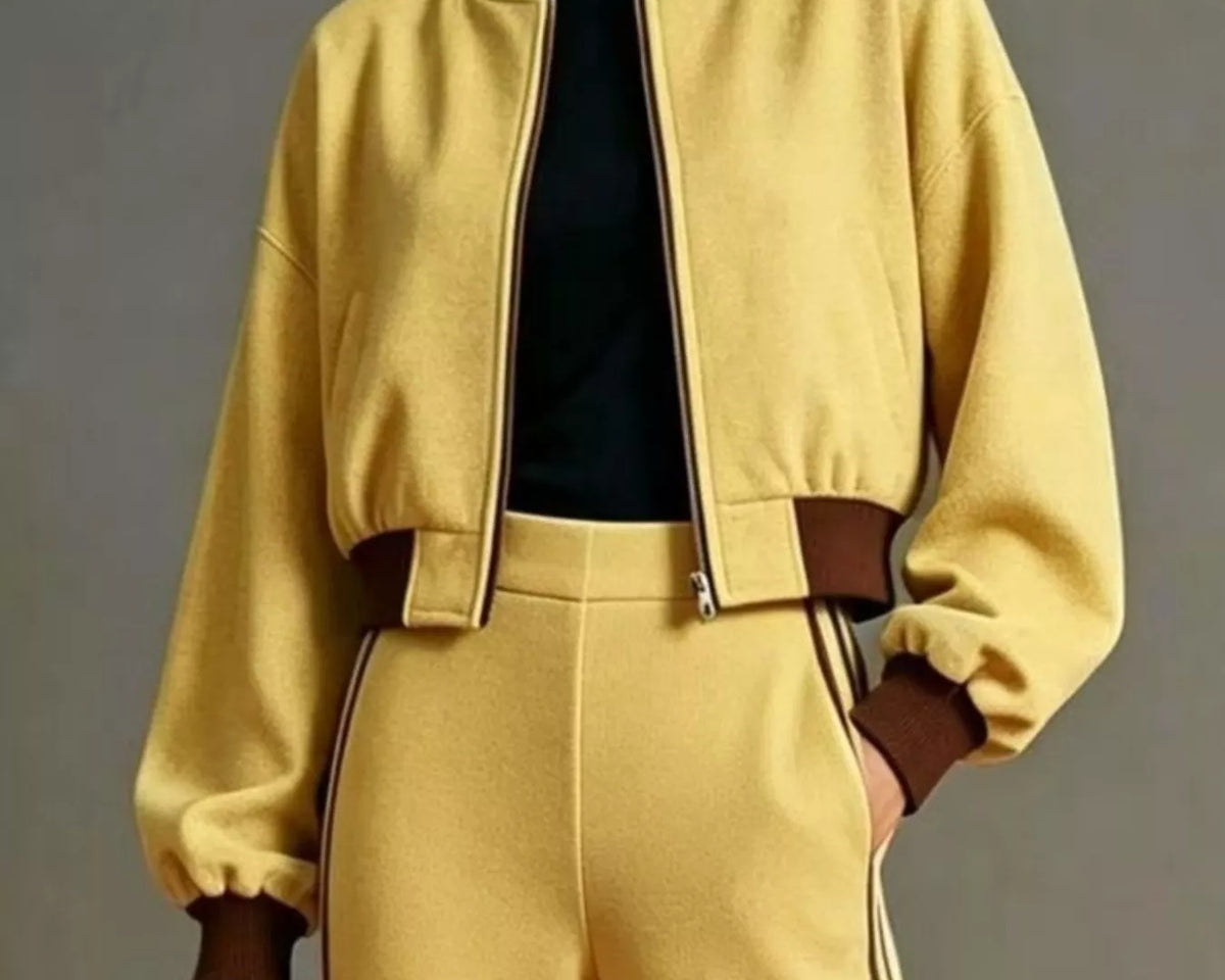 NEWYORKLISTED STYLE COLLECTION: A model wears a matching yellow tracksuit featuring a cropped zip-up bomber jacket with brown ribbed collar, cuffs, and waist detail, paired with matching yellow pants with brown side stripes and a black top underneath.