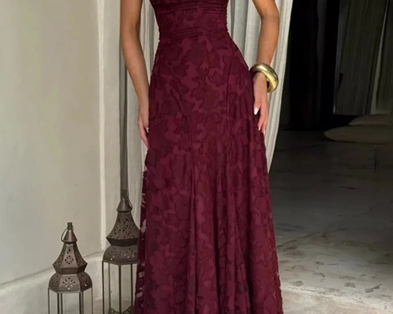 NEWYORKLISTED STYLE COLLECTION: Woman wearing a burgundy lace dress standing indoors with decorative lanterns and a plant in the background.