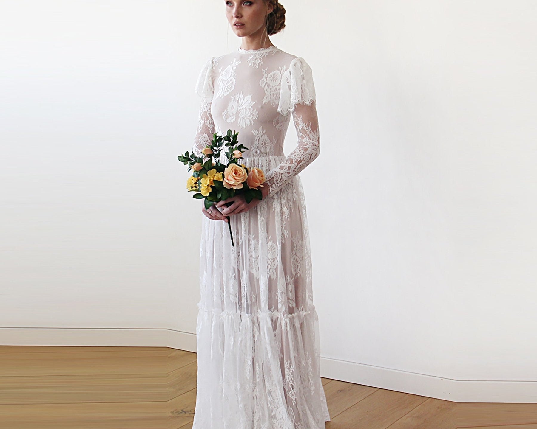 NEWYORKLISTED ELEGANT BRIDE COLLECTION: Woman in a white lace wedding dress holding a bouquet against a plain background