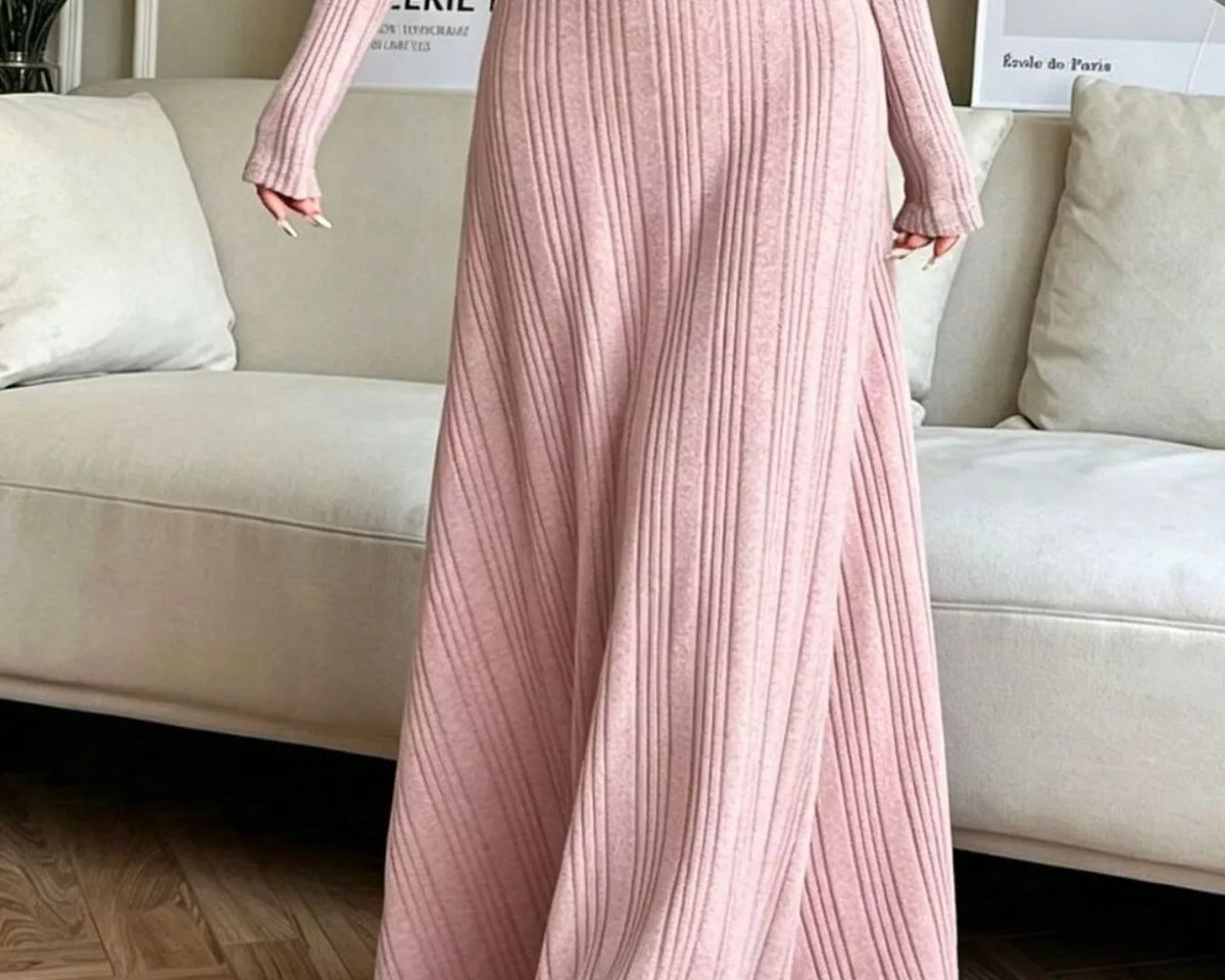 NEWYORKLISTED STYLE COLLECTION: A woman wearing a long-sleeve, ribbed knit maxi dress in a dusty pink color, standing in a living room setting with a beige sofa and wood flooring.