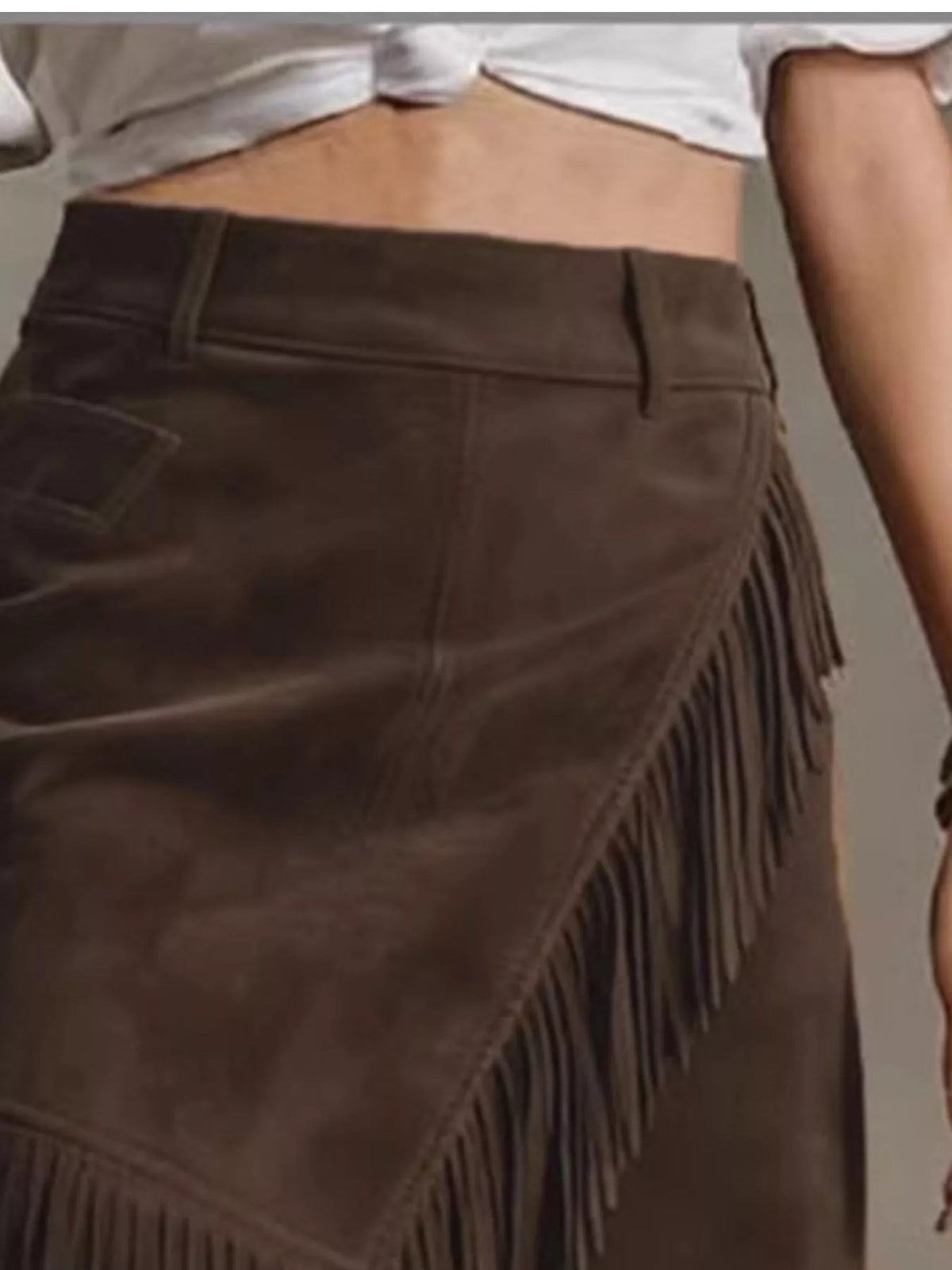 NEWYORKLISTED STYLE COLLECTION: A close-up of a dark brown suede skirt with diagonal fringe detailing, worn by a model in a cropped white top.