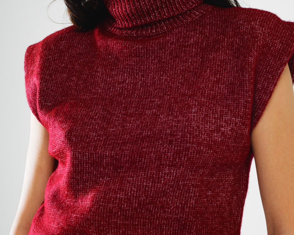 Woman wearing a red sleeveless knitted top with a belt, sunglasses, and a hat on a light gray background