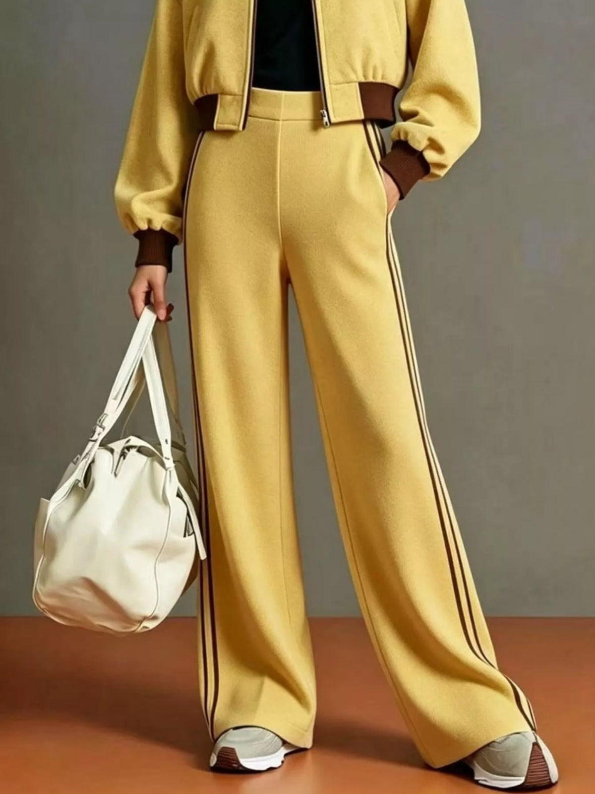 NEWYORKLISTED STYLE COLLECTION: A model wearing a matching yellow tracksuit with brown trim, featuring wide-leg pants with double dark stripes down the side, a cropped jacket, and sneakers, while holding a white shoulder bag against a gray studio background.
