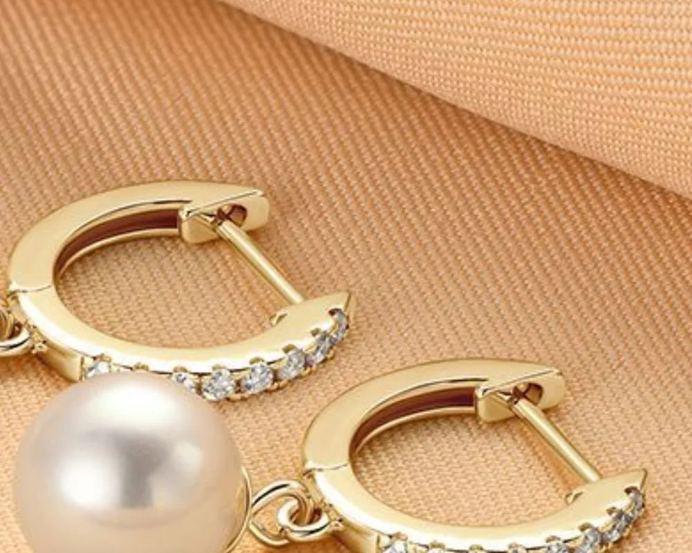 NYLD GLAMOUR COLLECTION by NEWYORKLISTED: Gold hoop earrings with a pearl and small diamonds on a beige fabric background
