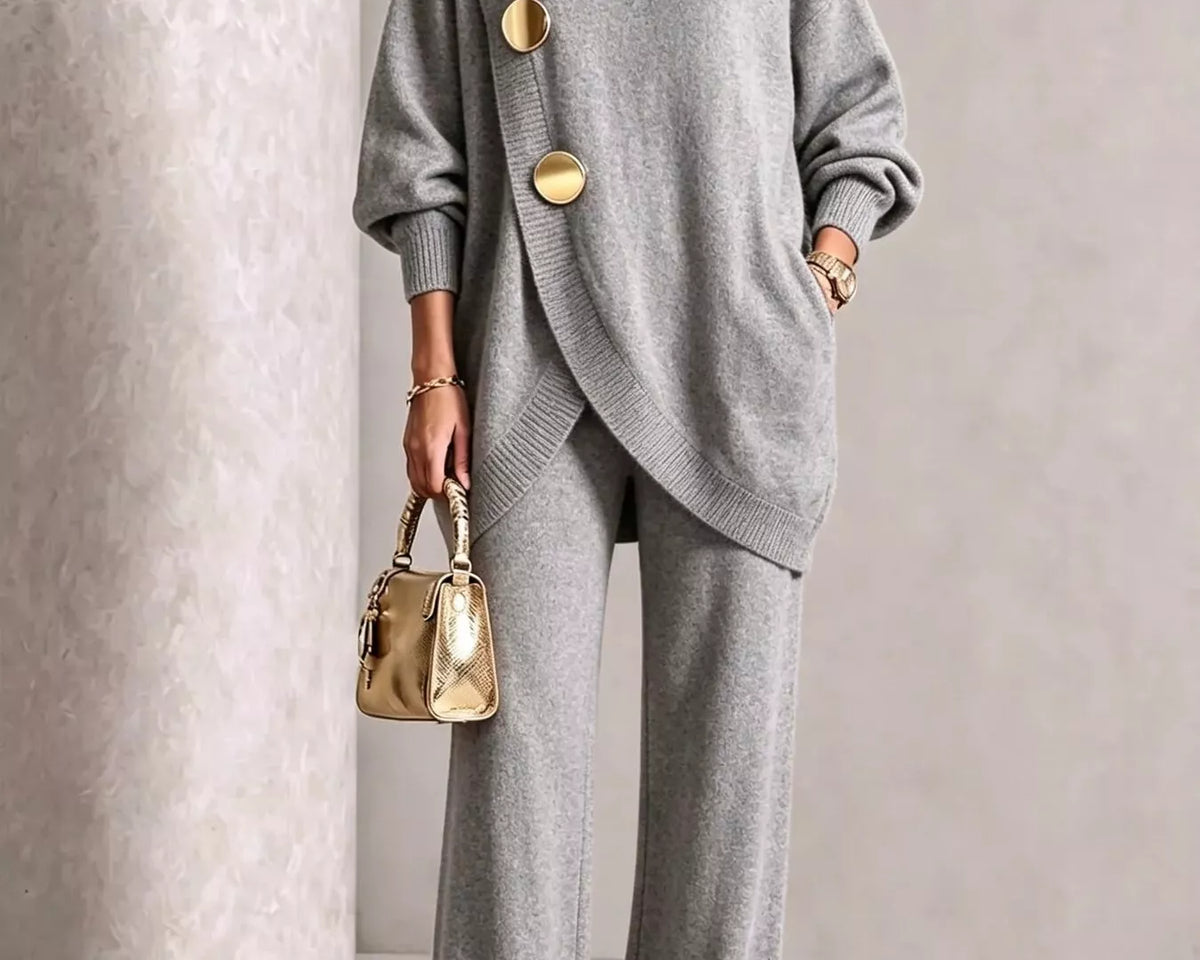 NEWYORKLISTED STYLE COLLECTION: A model wears a light gray knit two-piece set consisting of a turtleneck sweater with oversized sleeves and large gold-tone decorative buttons, paired with matching wide-leg trousers. She is holding a small metallic gold handbag and wearing gold pointed shoes, set against a textured light gray studio background.