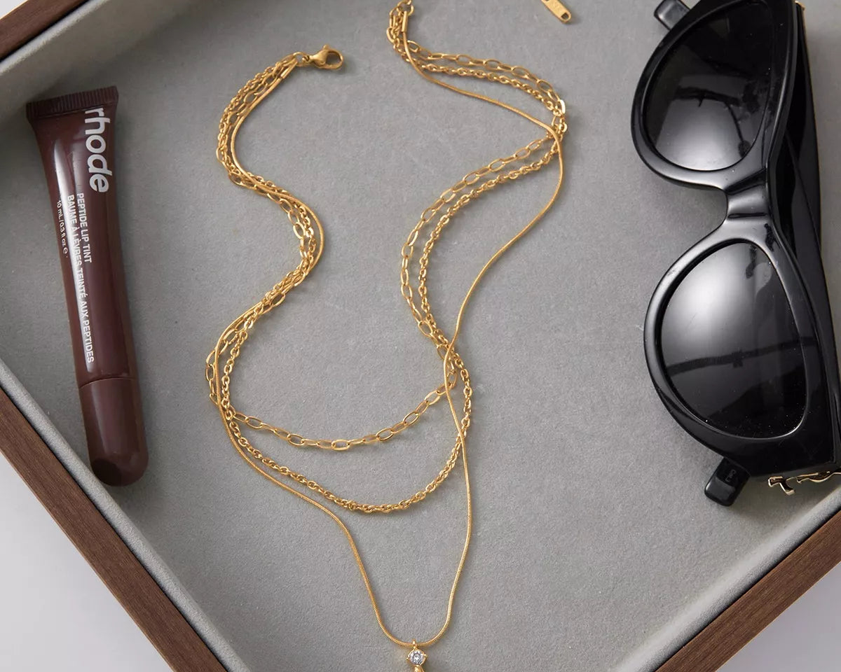 NYLD Glamour Collection by NEWYORKLISTED: Gold necklace with a pendant, tube of lipstick, and sunglasses on a gray surface.