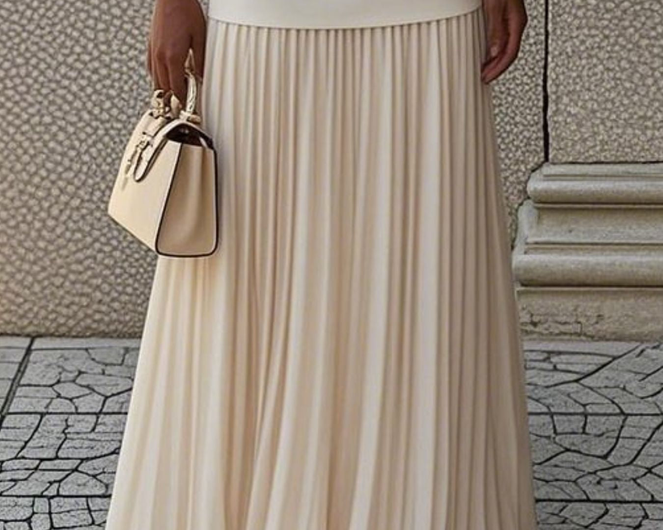 NEWYORKLISTED STYLE COLLECTION: Person wearing a beige pleated skirt holding a matching handbag on a stone pavement.