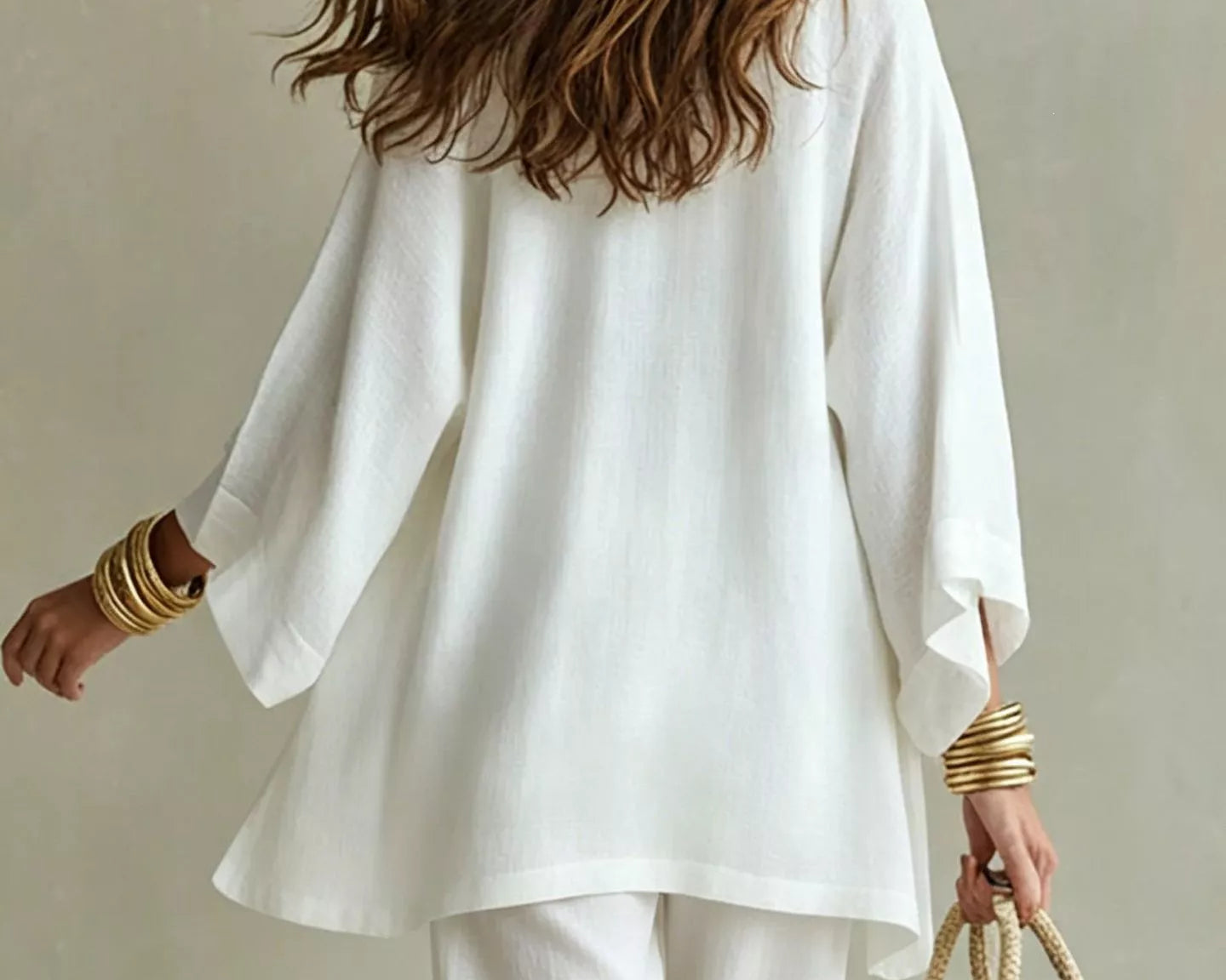 NEWYORKLISTED STYLE COLLECTION: A woman shown from behind wearing a loose white long-sleeve top and white pants, holding a woven straw tote bag against a plain beige background.