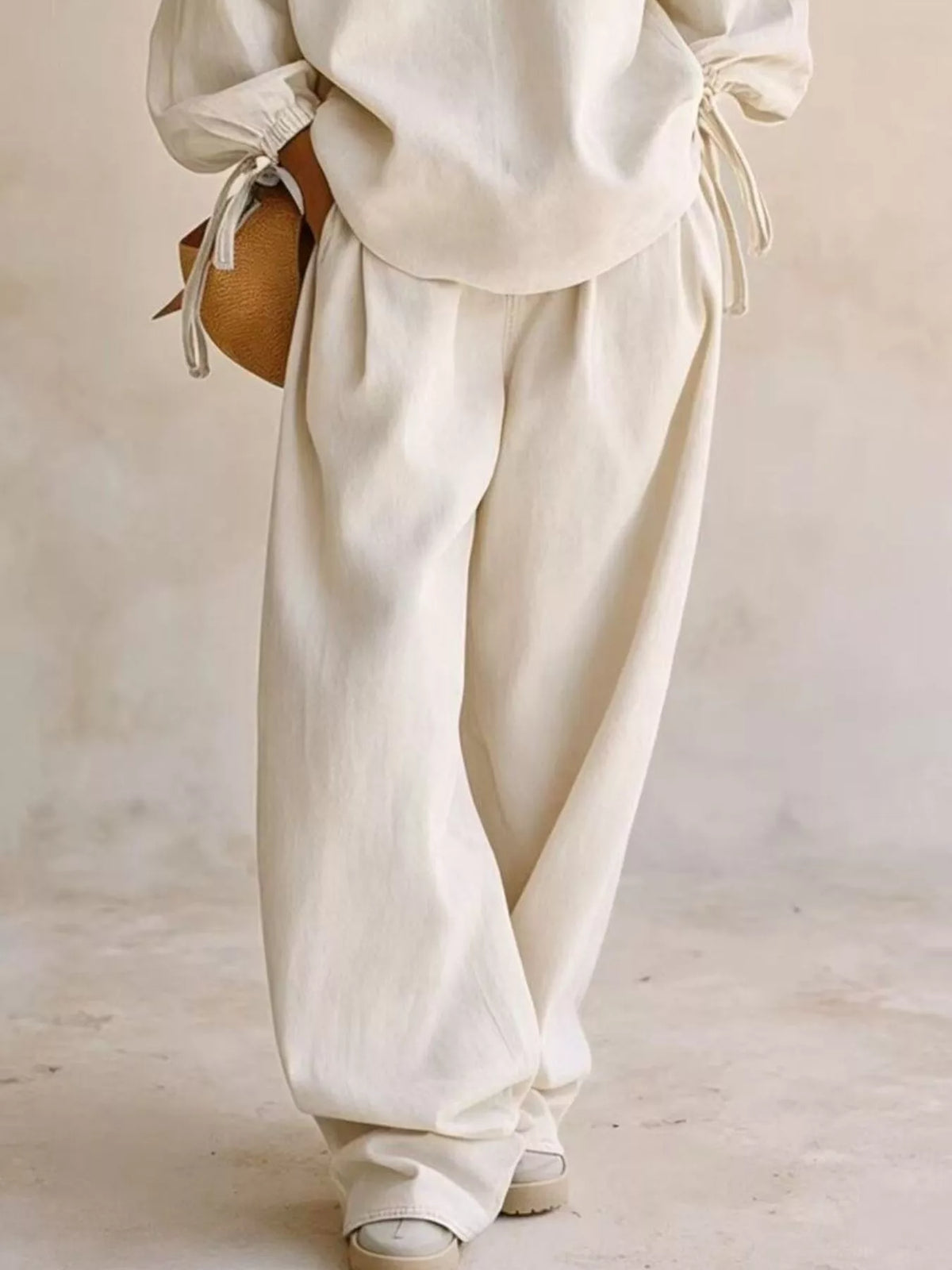 NEWYORKLISTED STYLE COLLECTION: A person wearing loose, wide-leg cream linen pants and a matching cream top, holding a small brown woven bag against a neutral, textured beige background.