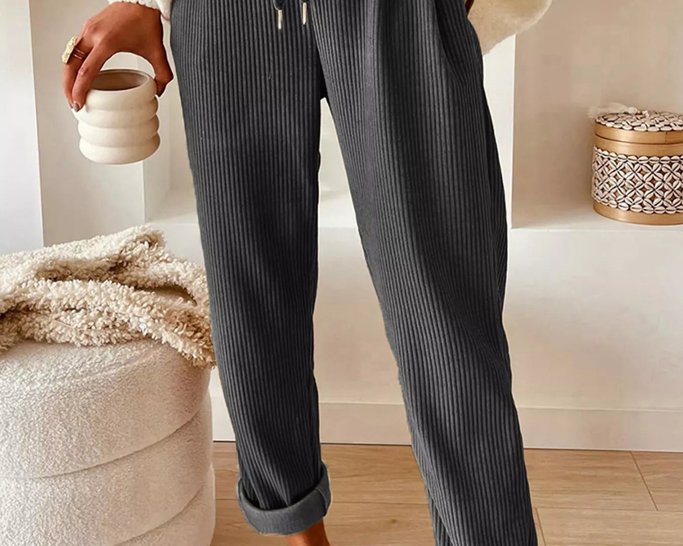 Person wearing dark ribbed pants with a white sweater and brown shoes, holding a ceramic cup.