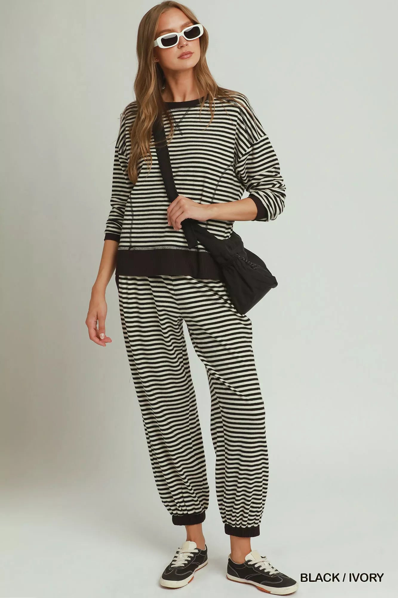 NEWYORKLISTED COMFORT COLLECTION: A woman modeling a matching black and ivory striped lounge set, featuring a long-sleeve top and jogger pants, styled with a black crossbody bag and black sneakers against a light gray studio background.