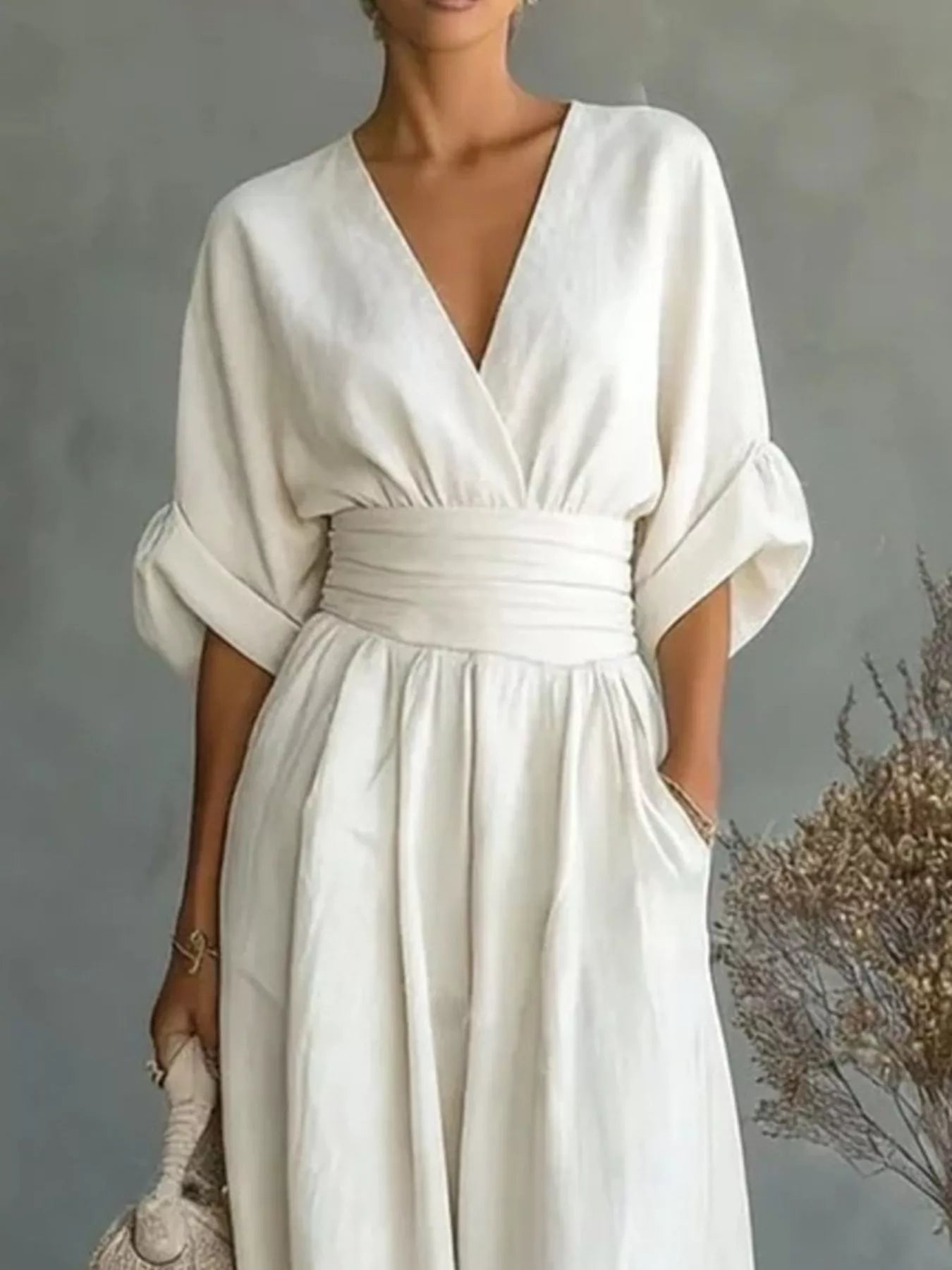 NEWYORKLISTED STYLE COLLECTION: A woman wearing a cream-colored linen jumpsuit with a deep V-neckline, wide three-quarter sleeves, and a matching fabric waist belt. She is holding a woven clutch against a textured gray background with dried floral decor.