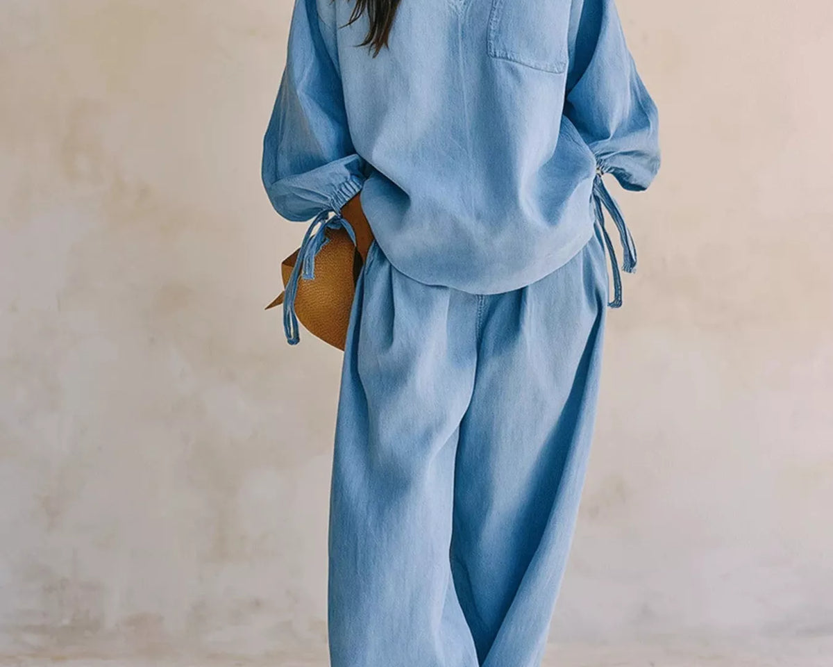 NEWYORKLISTED STYLE COLLECTION: A woman models a light blue linen two-piece set consisting of a long-sleeve V-neck top with tie details at the cuffs and matching wide-leg pants, set against a neutral textured beige wall.