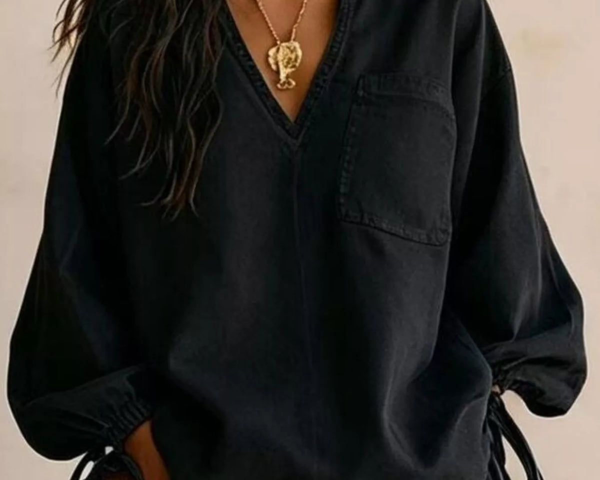 NEWYORKLISTED STYLE COLLECTION: A woman wearing an oversized black long-sleeve blouse with a split neckline, chest pocket, and side tie details, styled with gold jewelry and blue earrings against a neutral beige background.