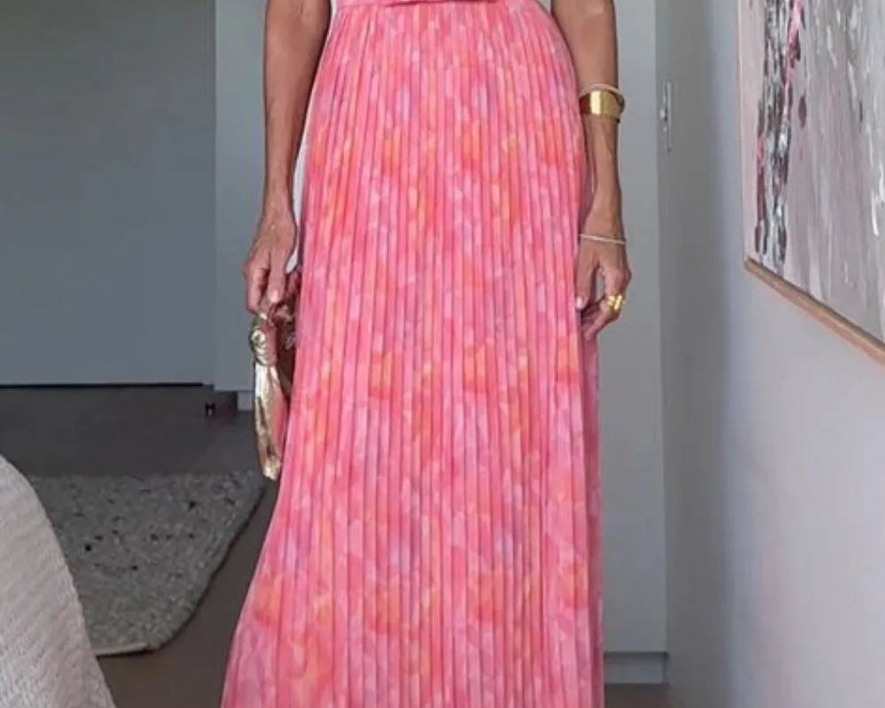 A flowing coral and pink halter neck pleated maxi dress with belted waist and backless design, perfect for summer weddings or resort evenings.