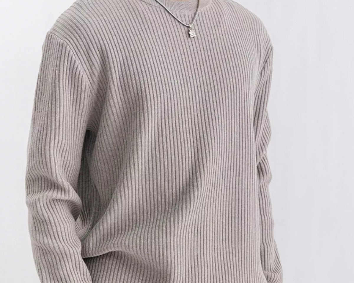 NEWYORKLISTED ATLAS MEN'S COLLECTION: A person wearing a light gray ribbed knit crewneck sweater, paired with tan pants and a silver necklace, photographed against a plain light gray background.