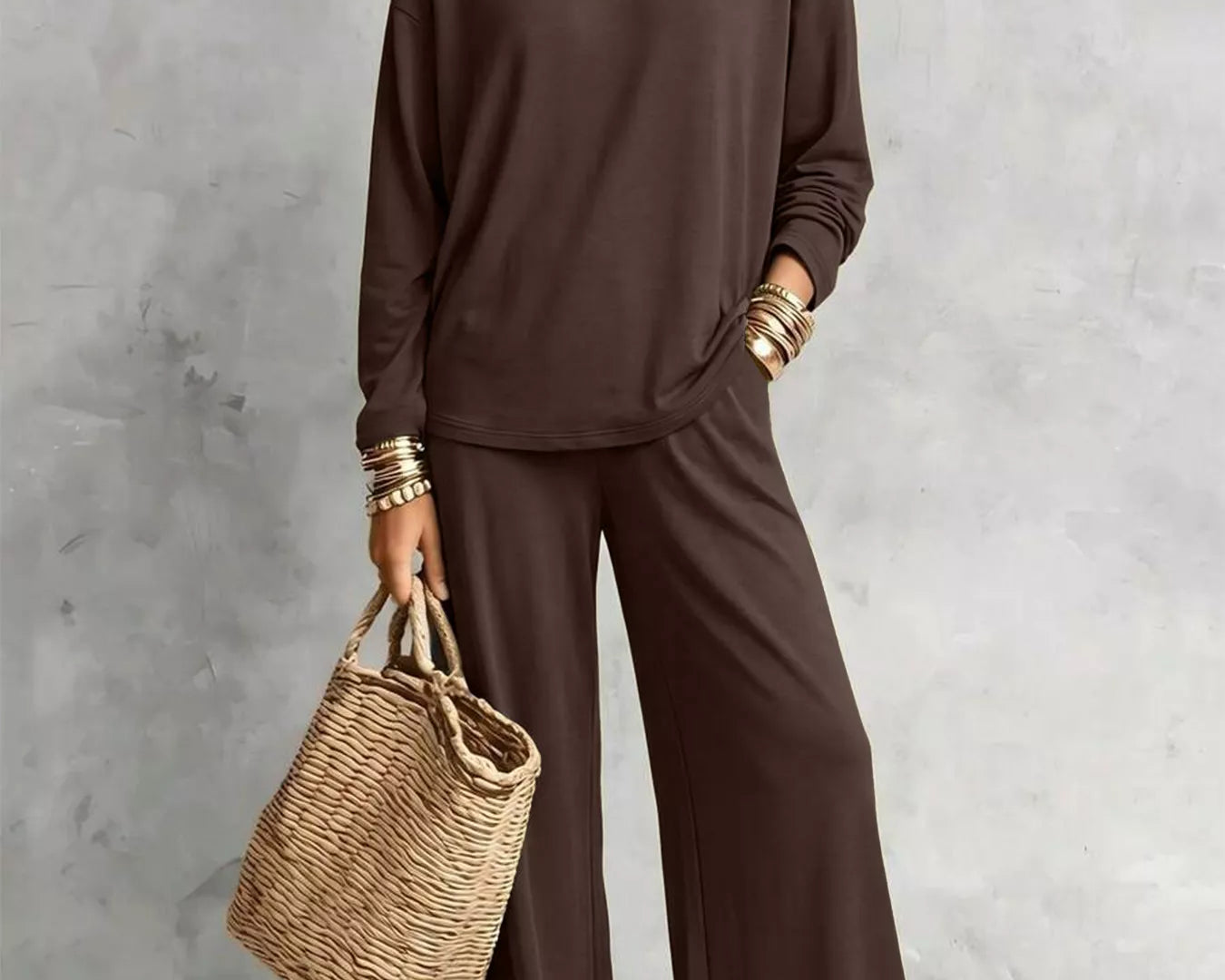 NEWYORKLISTED STYLE COLLECTION: A woman models a dark brown two-piece lounge set consisting of a long-sleeve top and matching wide-leg pants, styled with layered gold necklaces, bracelets, and a straw tote bag against a textured gray wall.