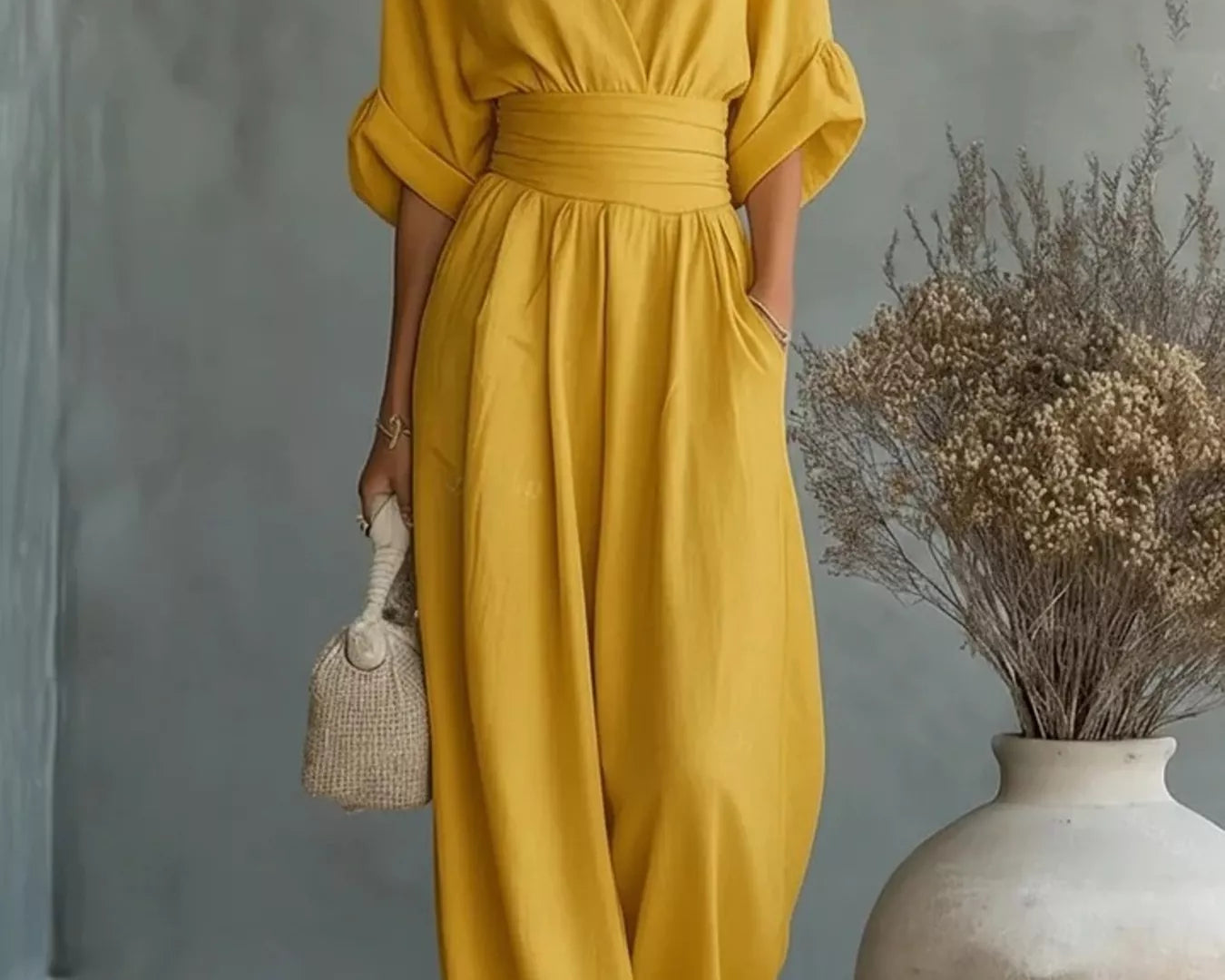 NEWYORKLISTED STYLE COLLECTION: Woman wearing a yellow jumpsuit standing next to a vase with dried flowers against a gray wall.