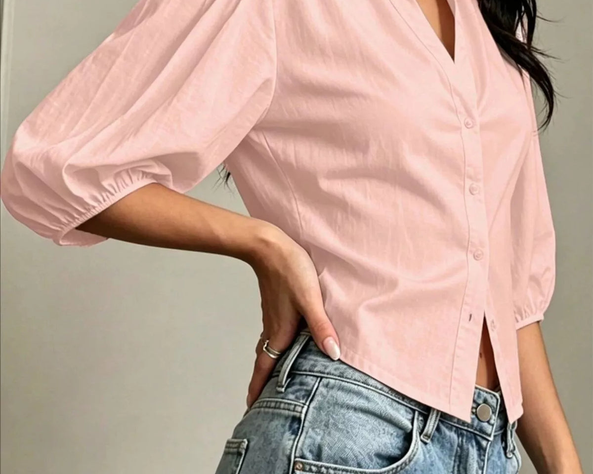 NEWYORKLISTED STYLE COLLECTION: A woman wearing a light pink button-up blouse with voluminous elbow-length puff sleeves and light-wash blue jeans, shown from the neck down against an indoor wall background.