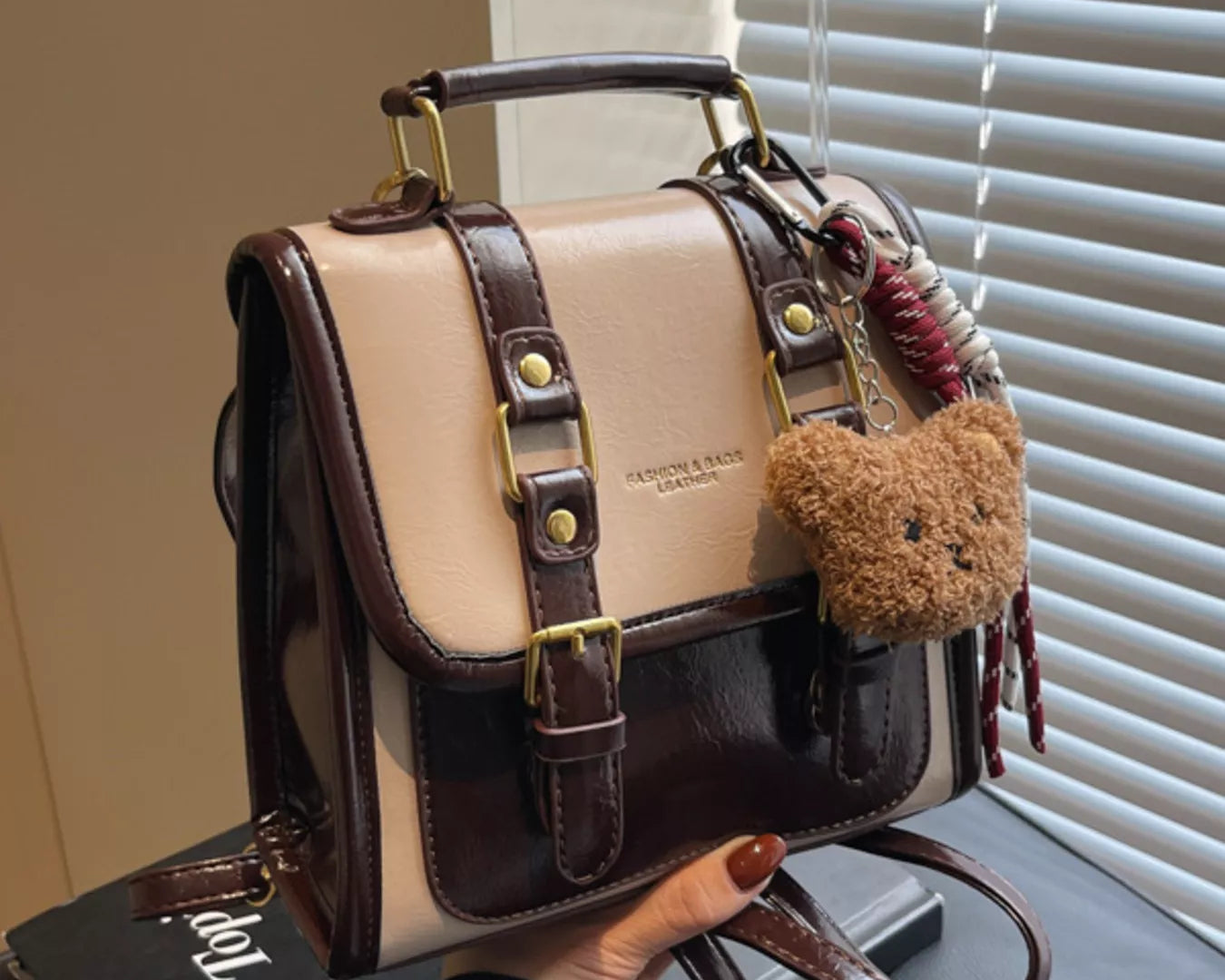NEWYORKLISTED STYLE COLLECTION: Beige and brown handbag with teddy bear charm held by a person, with a window and blinds in the background.