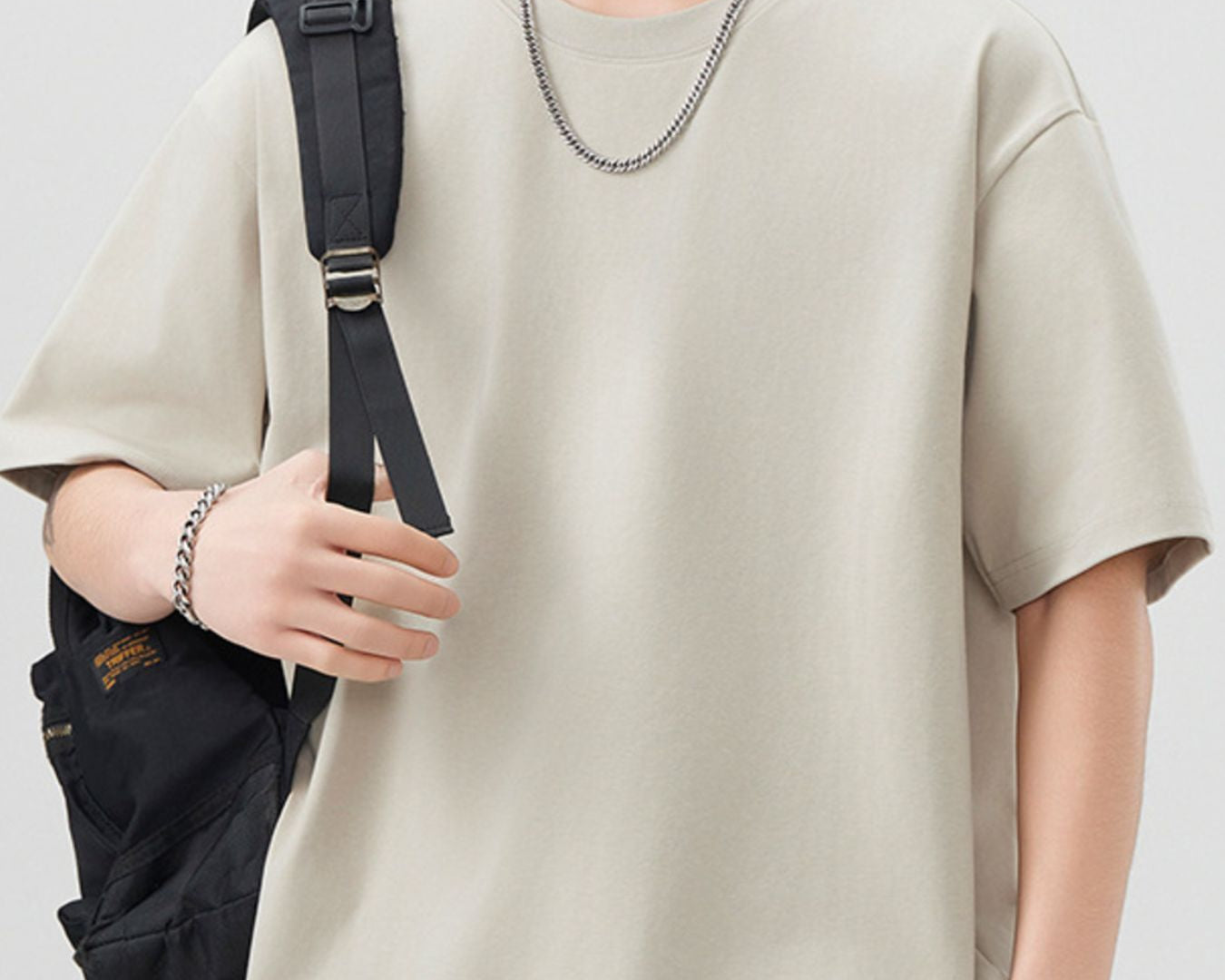 NEWYORKLISTED ATLAS MEN"S COLLECTION: Person wearing a beige t-shirt holding a black backpack against a white background