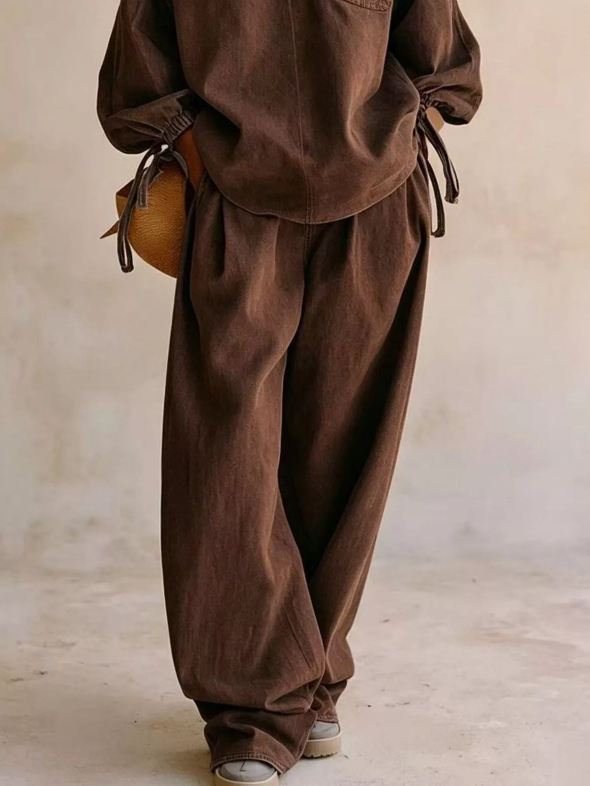 NEWYORKLISTED STYLE COLLECTION: A person wearing a matching brown linen-like two-piece outfit with a long-sleeve top and very loose wide-leg pants, standing against a neutral beige wall. A small tan woven bag is also visible.