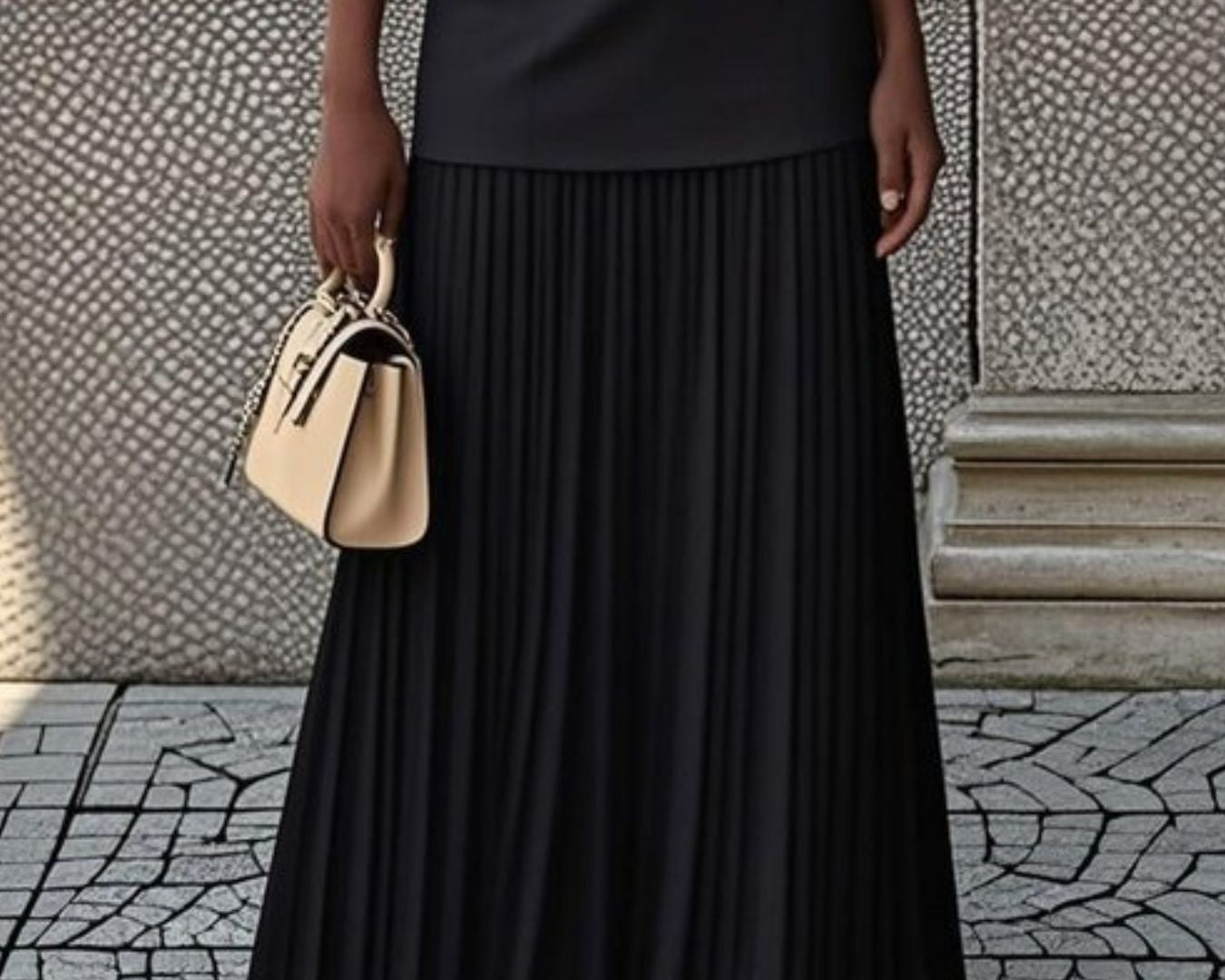 NEWYORKLISTED STYLE COLLECTION: Person wearing a black pleated skirt and holding a beige handbag against a textured wall.