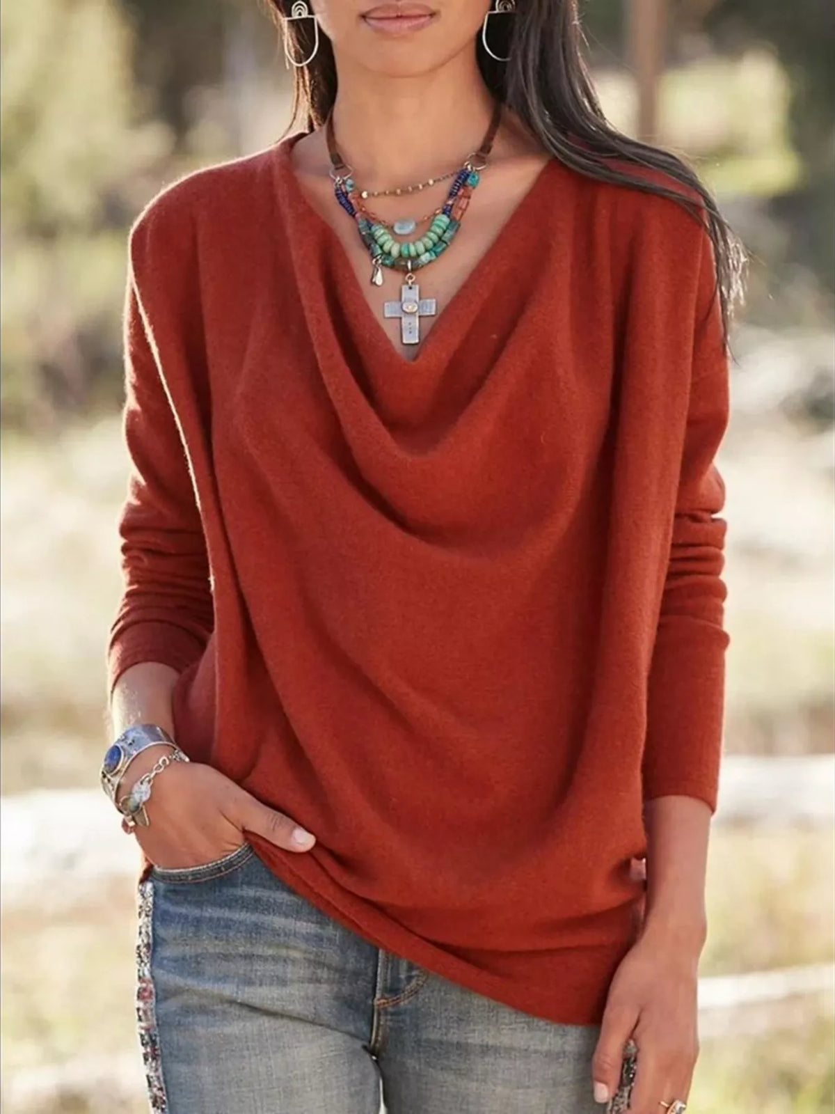 NEWYORKLISTED STYLE COLLECTION: A woman wearing a rust-orange long-sleeve knit top with a draped cowl neckline, paired with blue jeans and layered necklaces, photographed outdoors against a blurred natural background.