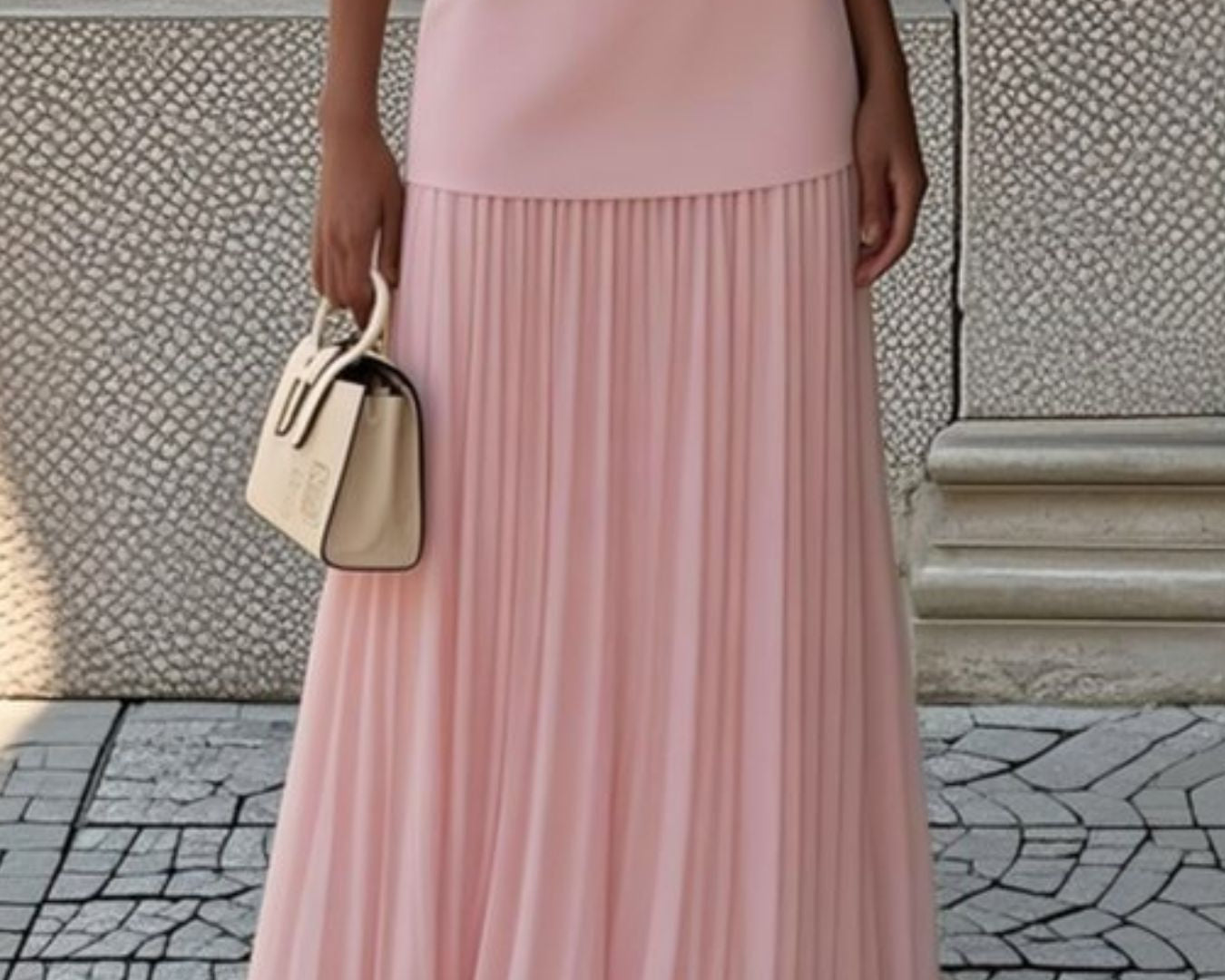 NEWYORKLISTED STYLE COLLECTION: Person wearing a pink pleated dress holding a beige handbag on a stone pavement.