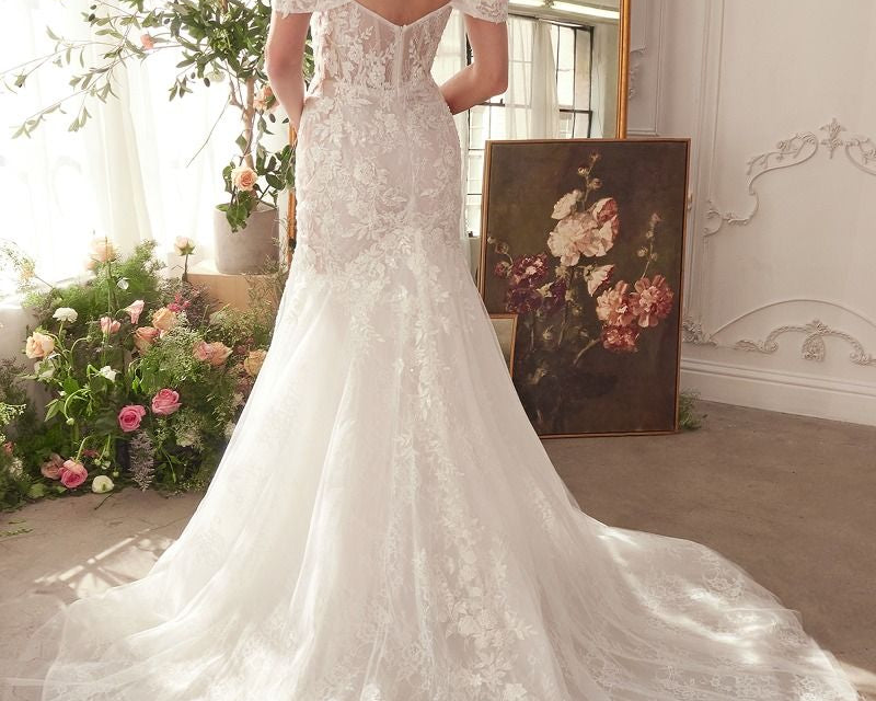 NEWYORKLISTED ELEGANT BRIDE COLLECTION: Woman in a white lace wedding dress standing in a decorated room with flowers and a mirror.