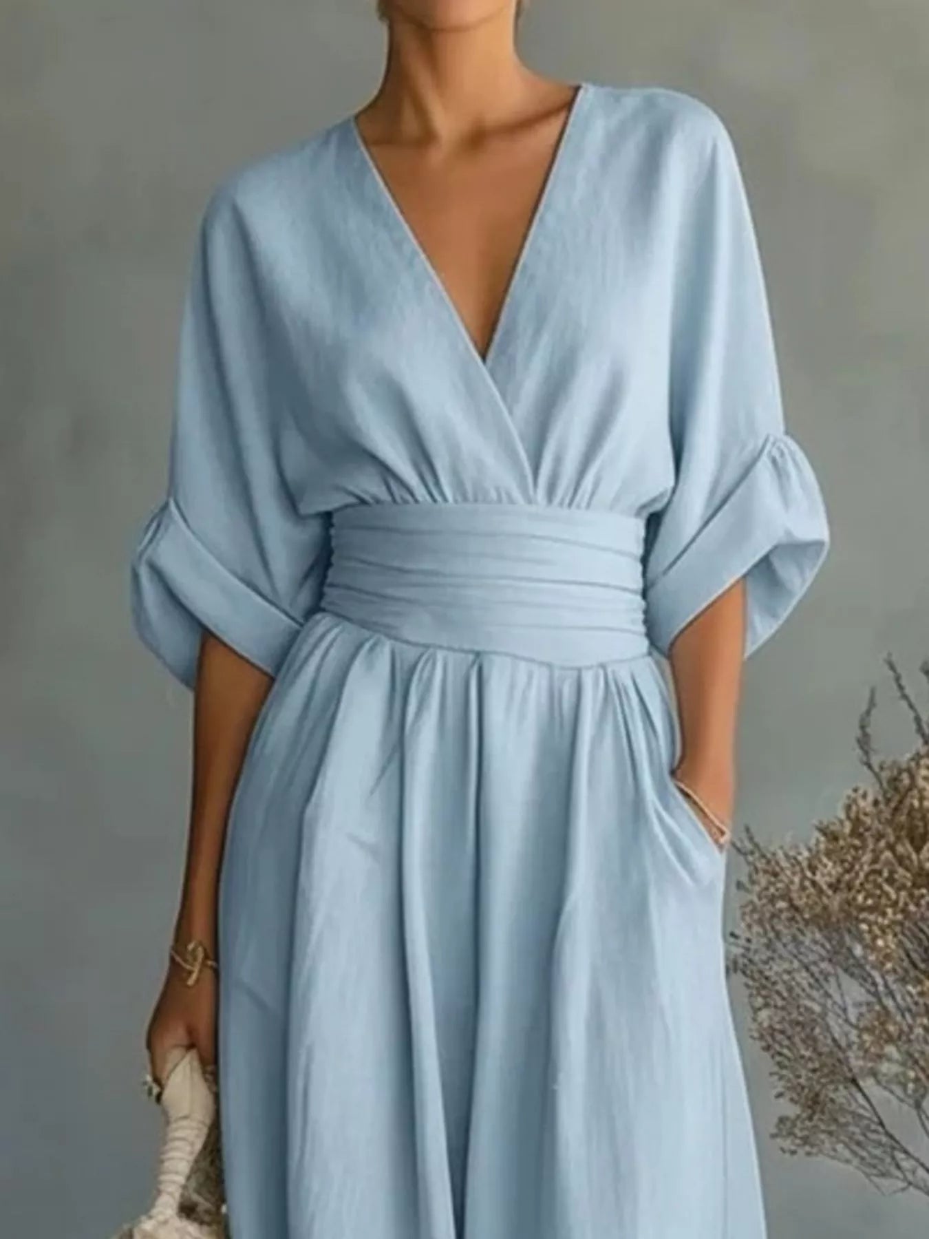 NEWYORKLISTED STYLE COLLECTION: A woman wearing a light blue midi dress with a wrap-style V-neckline, elbow-length puff sleeves, a matching fabric waist tie, and side pockets, photographed against a soft gray wall with dried floral decor.