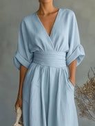 NEWYORKLISTED STYLE COLLECTION: A woman wearing a light blue midi dress with a wrap-style V-neckline, elbow-length puff sleeves, a matching fabric waist tie, and side pockets, photographed against a soft gray wall with dried floral decor.