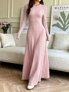 NEWYORKLISTED STYLE COLLECTION: A woman wearing a long-sleeve, floor-length pink ribbed knit dress, standing in a living room setting with a beige sofa and framed wall art in the background.