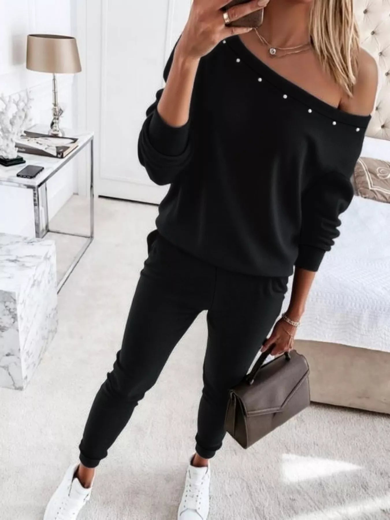 NEWYORKLISTED STYLE COLLECTION: Person wearing a black off-shoulder top with pearl details and black jogger pants, holding a brown handbag in a modern living room.
