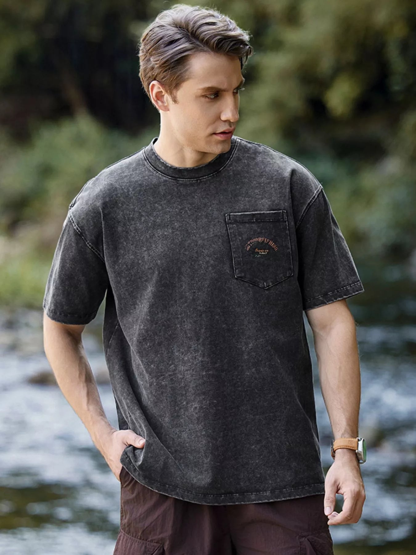 NEWYORKLISTED ATLAS MEN's COLLECTION: Man wearing a black t-shirt with a pocket, standing by a river with greenery in the background