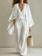 NEWYORKLISTED STYLE COLLECTION: A woman models a white three-piece linen outfit consisting of a V-neck knit top, matching wide-leg trousers with a drawstring, and a draped open-front cardigan. She accessorizes with layered gold necklaces, bracelets, and a woven straw handbag against a neutral beige background.
