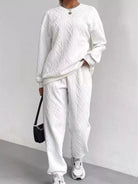 NEWYORKLISTED COMFORT COLLECTION: Person wearing a white cable knit sweater and pants with sneakers, holding a black bag against a light gray background.