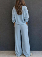 NEWYORKLISTED STYLE & COMFORT COLLECTION: A woman viewed from behind wearing a light blue knit matching set consisting of a long-sleeve top and wide-leg pants, standing against a dark gray textured wall.