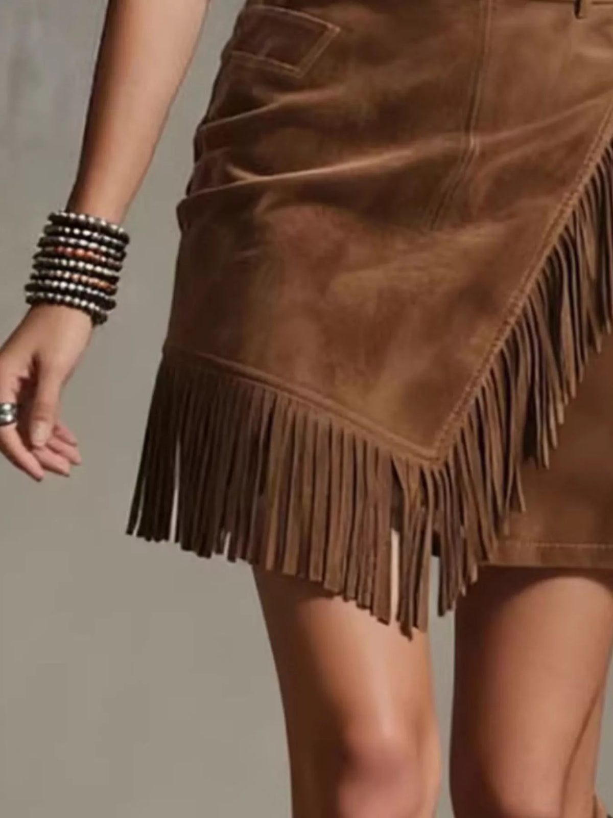 NEWYORKLISTED STYLE COLLECTION: A close-up of a brown suede fringe mini skirt worn by a model, showing the textured suede material, asymmetrical fringe hem, and side pockets.