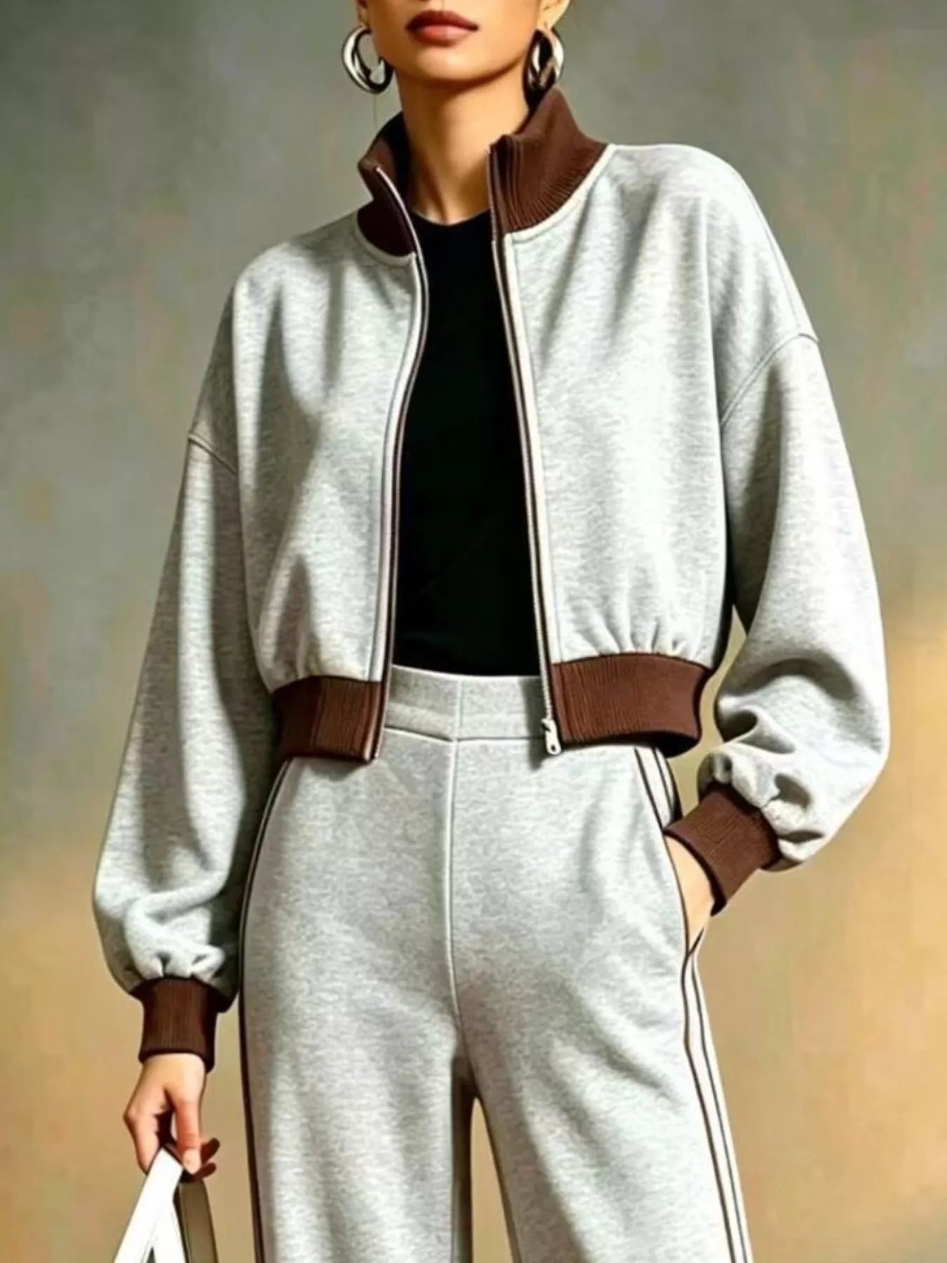 NEWYORKLISTED STYLE COLLECTION:  A person wearing a light gray cropped zip-up jacket with brown ribbed collar, hem, and cuffs, paired with matching light gray sweatpants and a black top, against a soft neutral studio background.