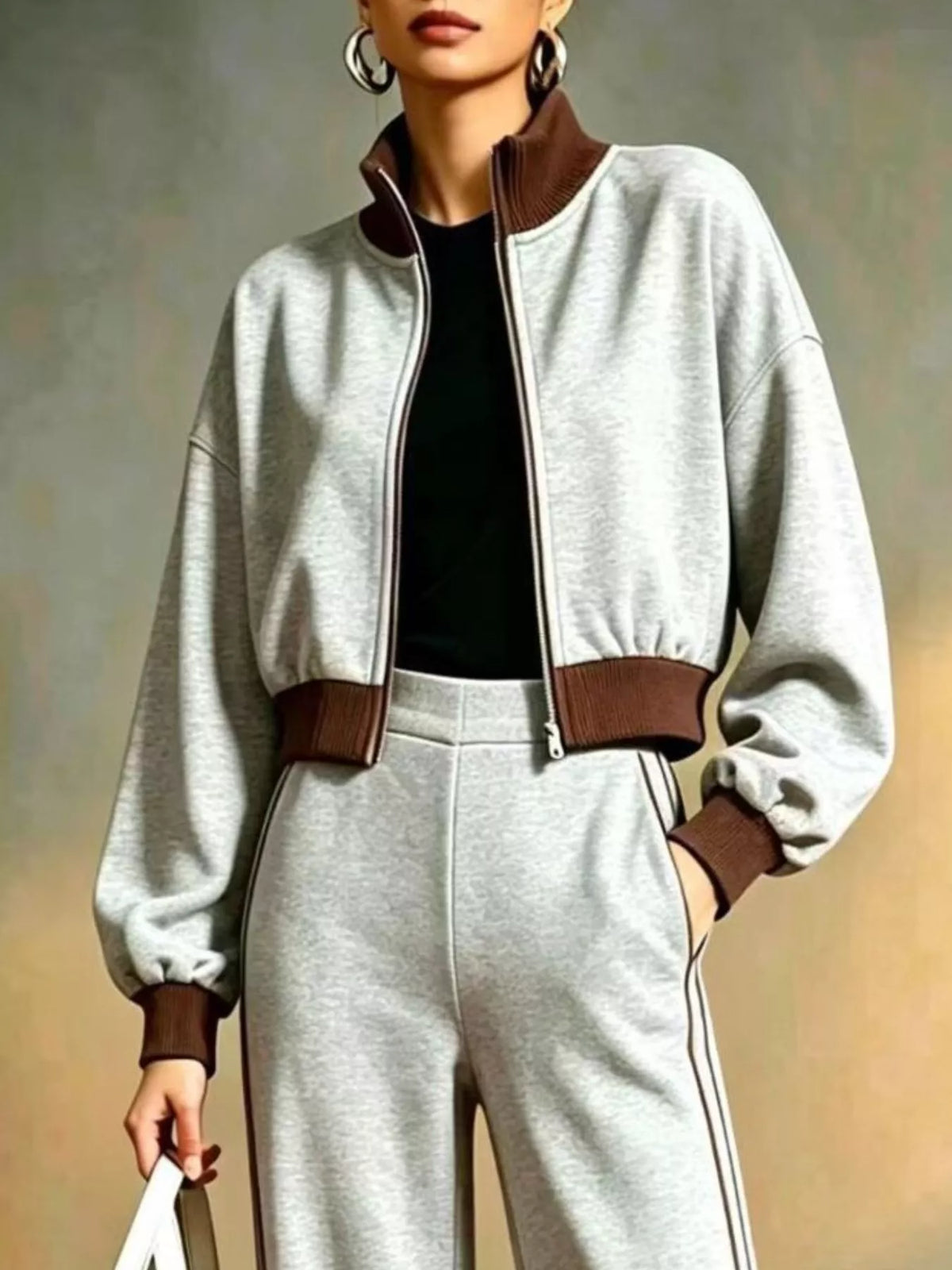 NEWYORKLISTED STYLE COLLECTION:  A person wearing a light gray cropped zip-up jacket with brown ribbed collar, hem, and cuffs, paired with matching light gray sweatpants and a black top, against a soft neutral studio background.