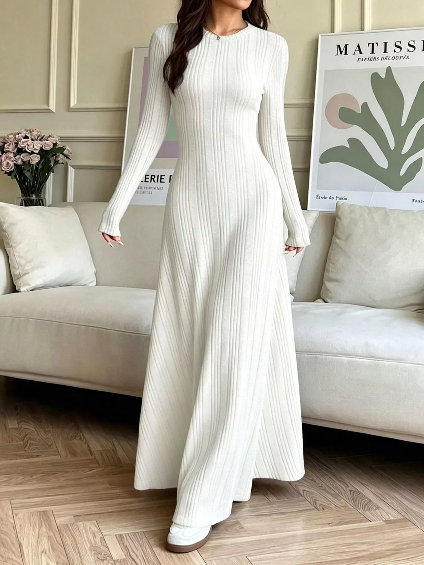 NEWYORKLISTED STYLE COLLECTION: A woman wearing a long-sleeve, floor-length white ribbed knit dress, standing in a living room setting with a beige sofa and framed wall art in the background.