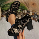 NEWYORKLISTED: Black velvet shoes with gold floral patterns held by a person against a natural background.