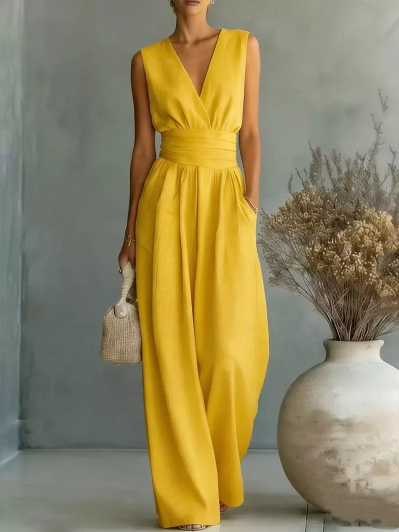 NEWYORKLISTED STYLE COLLECTION: Woman wearing a yellow jumpsuit standing next to a vase with dried plants against a gray wall.