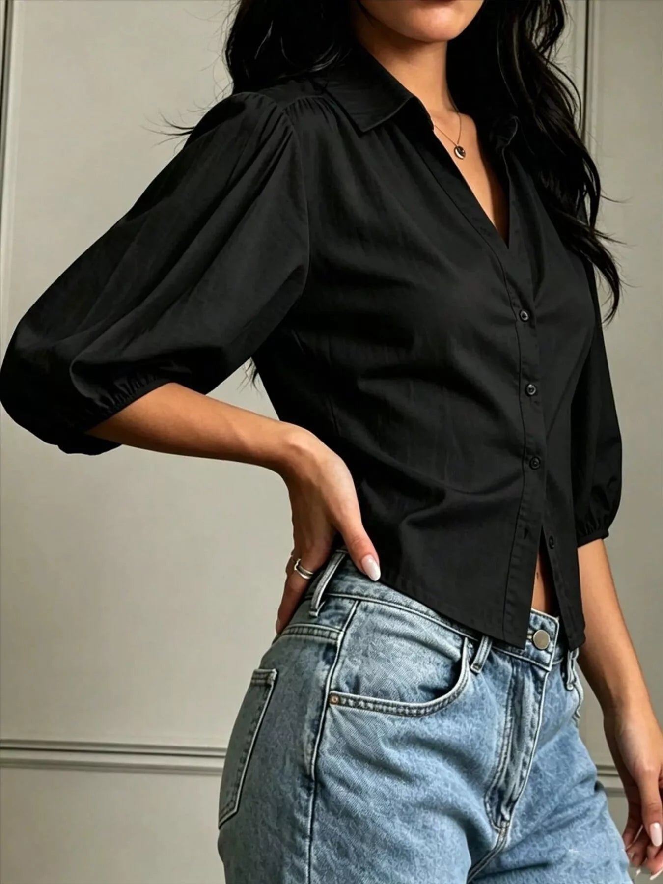 NEWYORKLISTED STYLE COLLECTION: A woman wearing a black button-up blouse with voluminous elbow-length puff sleeves and light-wash blue jeans, shown from the neck down against a neutral indoor wall.