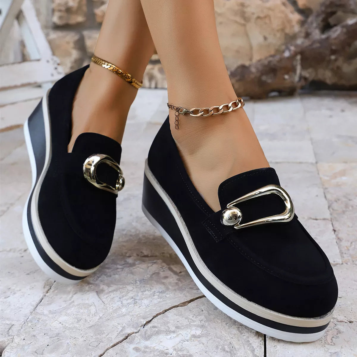 NEWYORKLISTED SHOE COLLECTION: Black loafers with gold buckles worn by a person on a light stone floor.