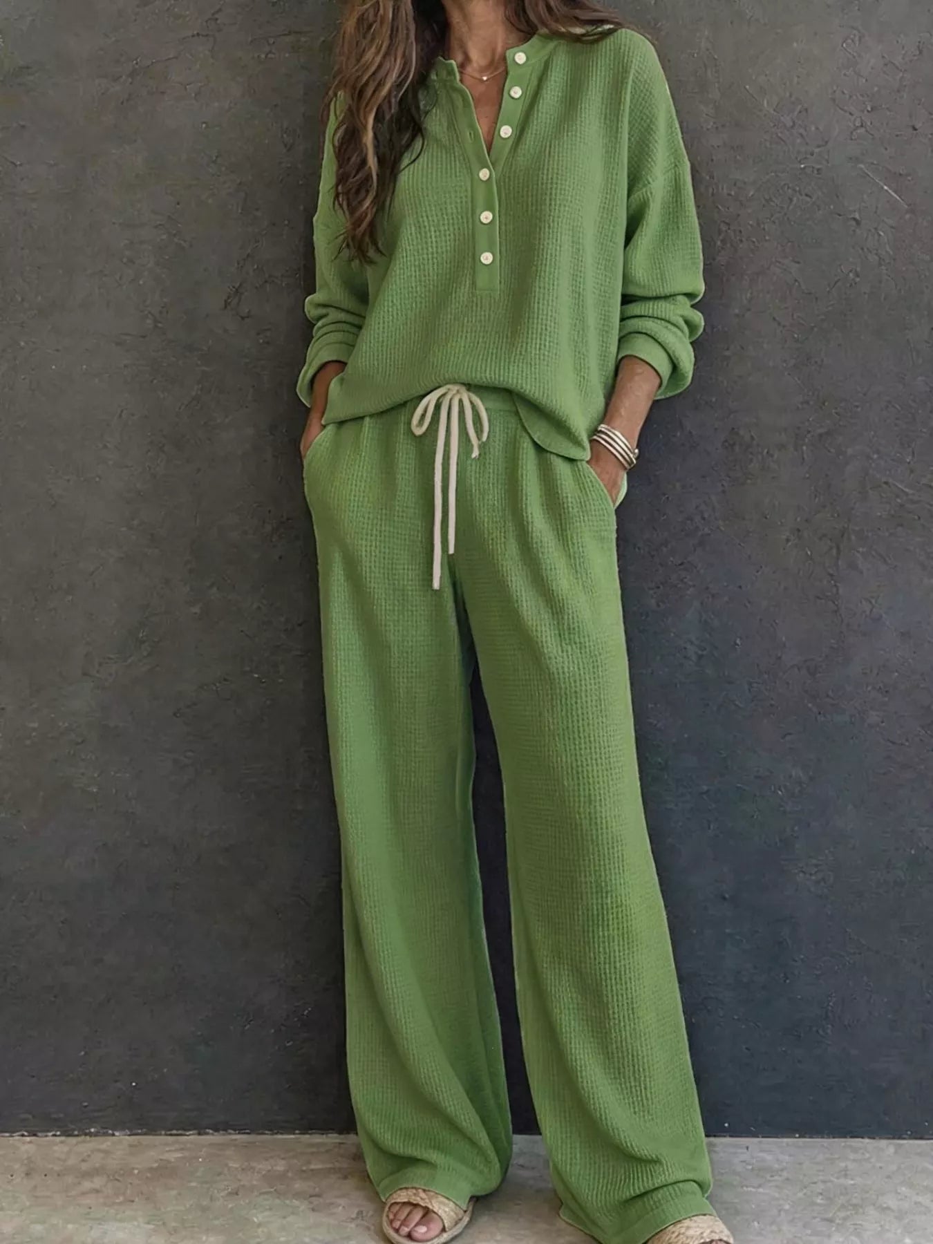 NEWYORKLISTED STYLE & COMFORT COLLECTION: A woman wearing a matching sage green knit lounge set consisting of a long-sleeve button-front top and wide-leg pants with a drawstring waist, standing against a dark textured wall.