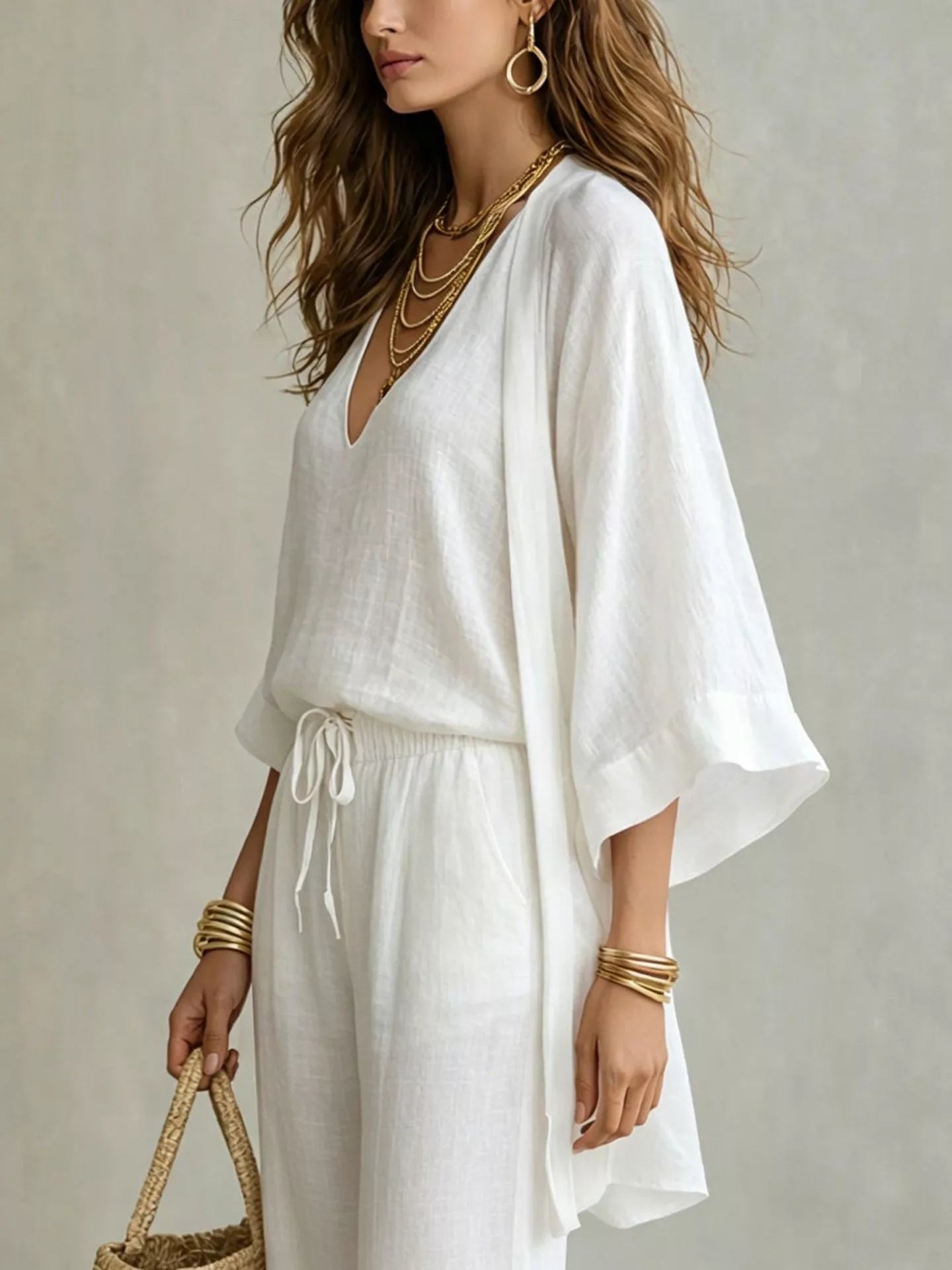 NEWYORKLISTED STYLE COLLECTION: A woman wearing a white linen kaftan-style dress with wide bell sleeves, a V-neckline, and a drawstring waist, styled with layered gold necklaces, bracelets, hoop earrings, and a woven straw handbag against a neutral textured background.